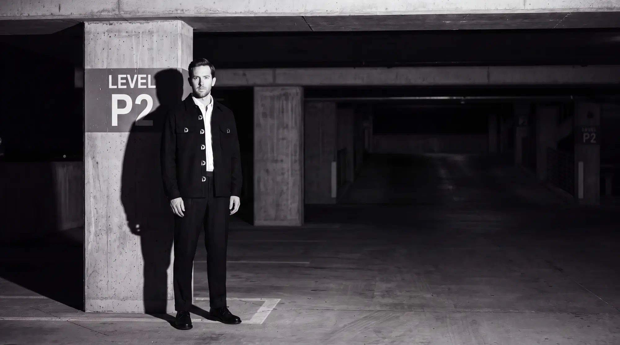 A man in a black wool overshirt, white oxford shirt and black trousers standing in an underground parking garage at night, lit by flash.