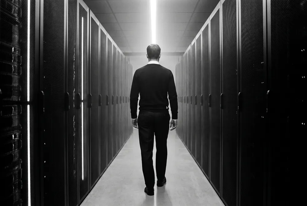 A man walking down a data-hall aisle between server racks in the black Merino Crewneck over a white Oxford Shirt.