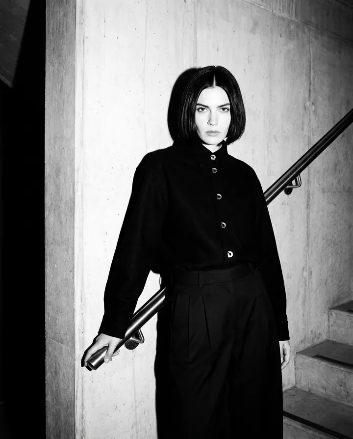 A woman in the black Melton Overshirt and Pleated Trouser on a concrete stairwell, lit by flash.